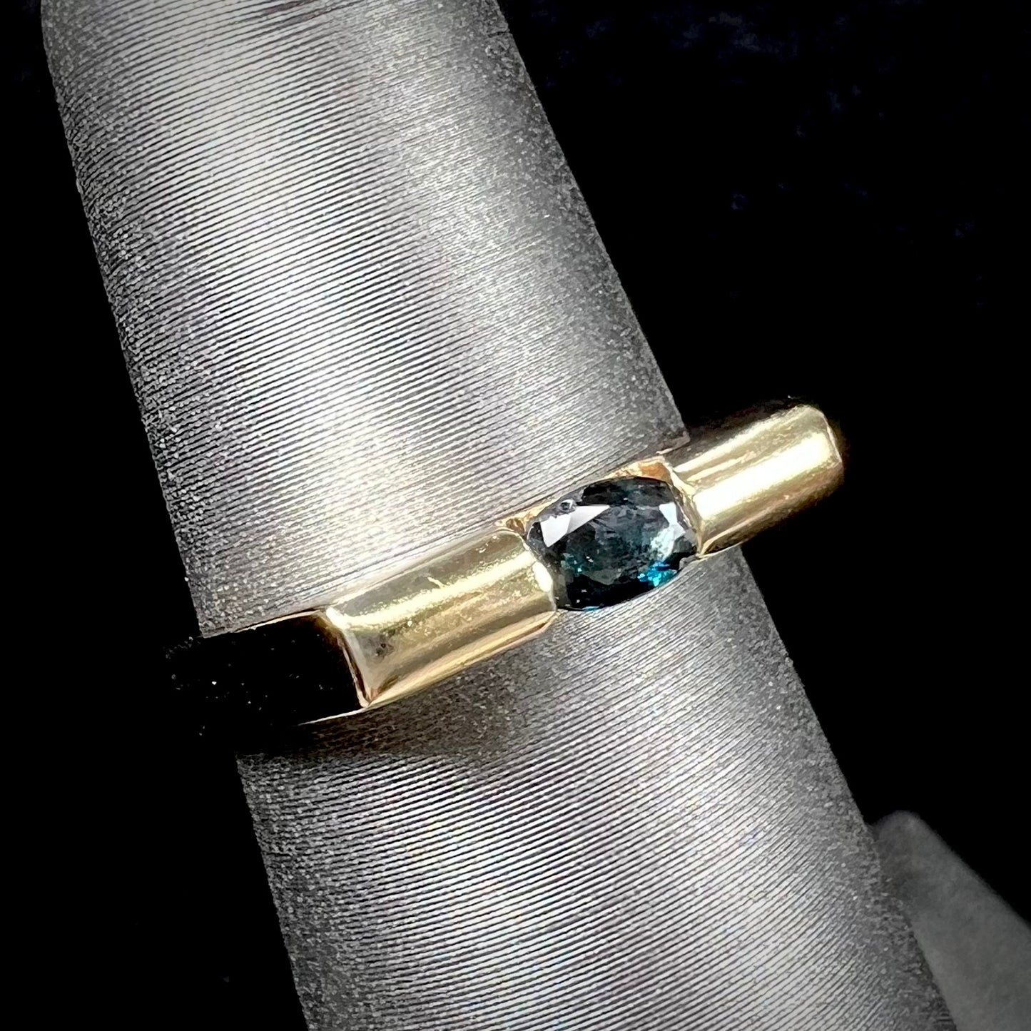 A ladies' yellow gold solitire ring horizontally set with an oval cut dark teal blue sapphire stone.