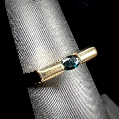 A ladies' yellow gold solitire ring horizontally set with an oval cut dark teal blue sapphire stone.