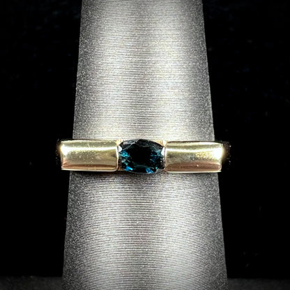 A ladies' yellow gold solitire ring horizontally set with an oval cut dark teal blue sapphire stone.