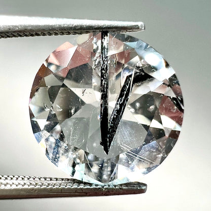 A round brilliant cut clear quartz stone with two black tourmaline crystal inclusions.