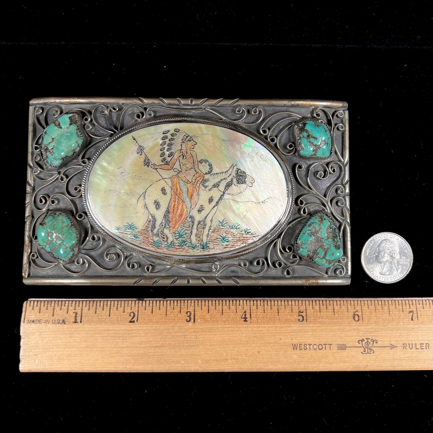 A men's Navajo-made white brass belt buckle set with turquoise nuggets and a mother of pearl stone etched with a Native American riding a horse.