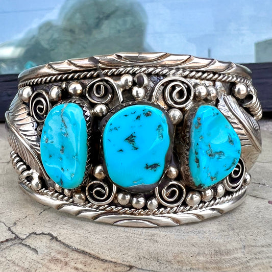 An ornate men's sterling silver turquoise cuff bracelet.  There are three turquoise stones set with silver feathers.