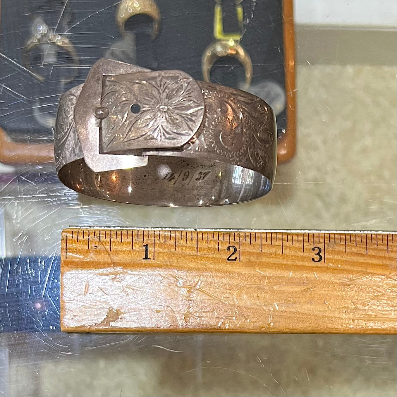 A photo showing a sterling silver women's antique Art Nouveau buckle bracelet.  The bracelet is engraved and etched with a floral pattern design.  The piece is being compared to a wooden ruler and measures 2.5-2.75 inches.