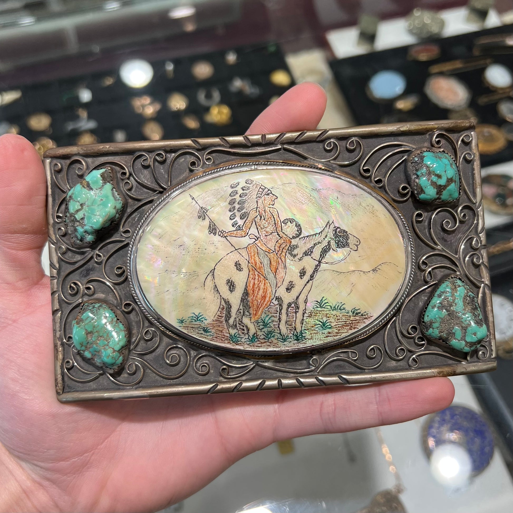 A men's Navajo-made white brass belt buckle set with turquoise nuggets and a mother of pearl stone etched with a Native American riding a horse.