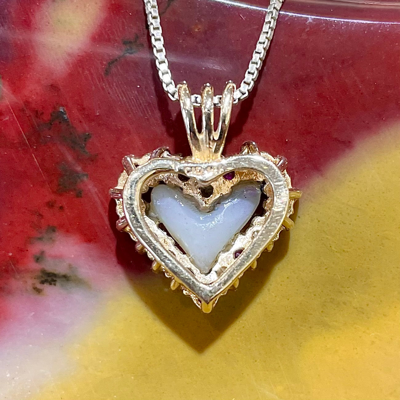A yellow gold pendant set with a heart cut white opal in a halo of alternating round rubies and diamonds.