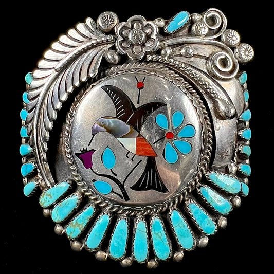 A sterling silver cuff bracelet featuring the motif of a stone inlaid hummingbird with petit point turquoise accents, handmade by Navajo artist Leo G. Harvey.