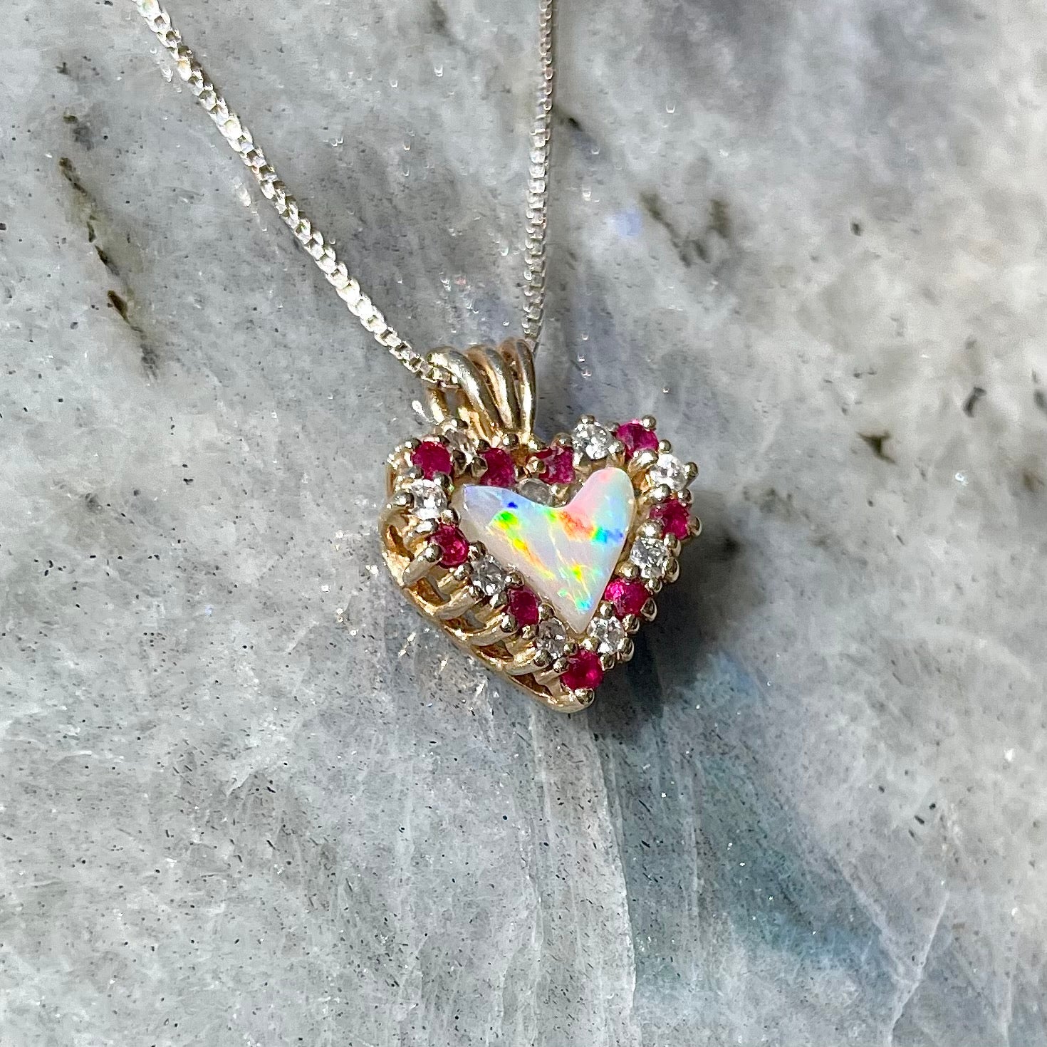 A yellow gold pendant set with a heart cut white opal in a halo of alternating round rubies and diamonds.