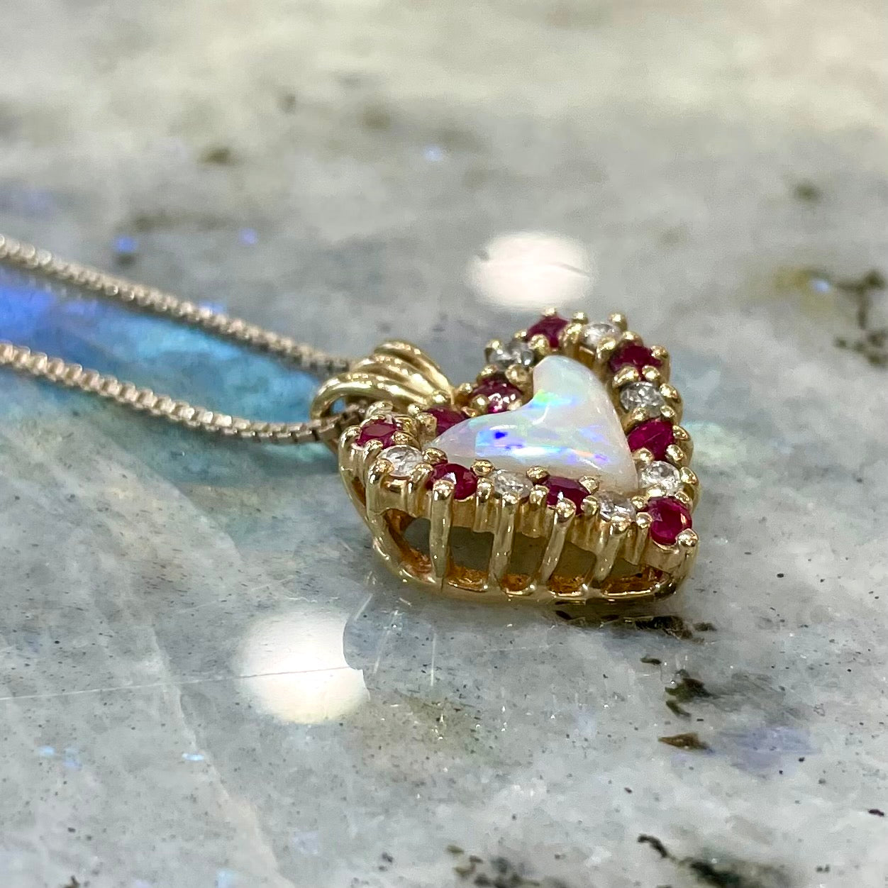 A yellow gold pendant set with a heart cut white opal in a halo of alternating round rubies and diamonds.