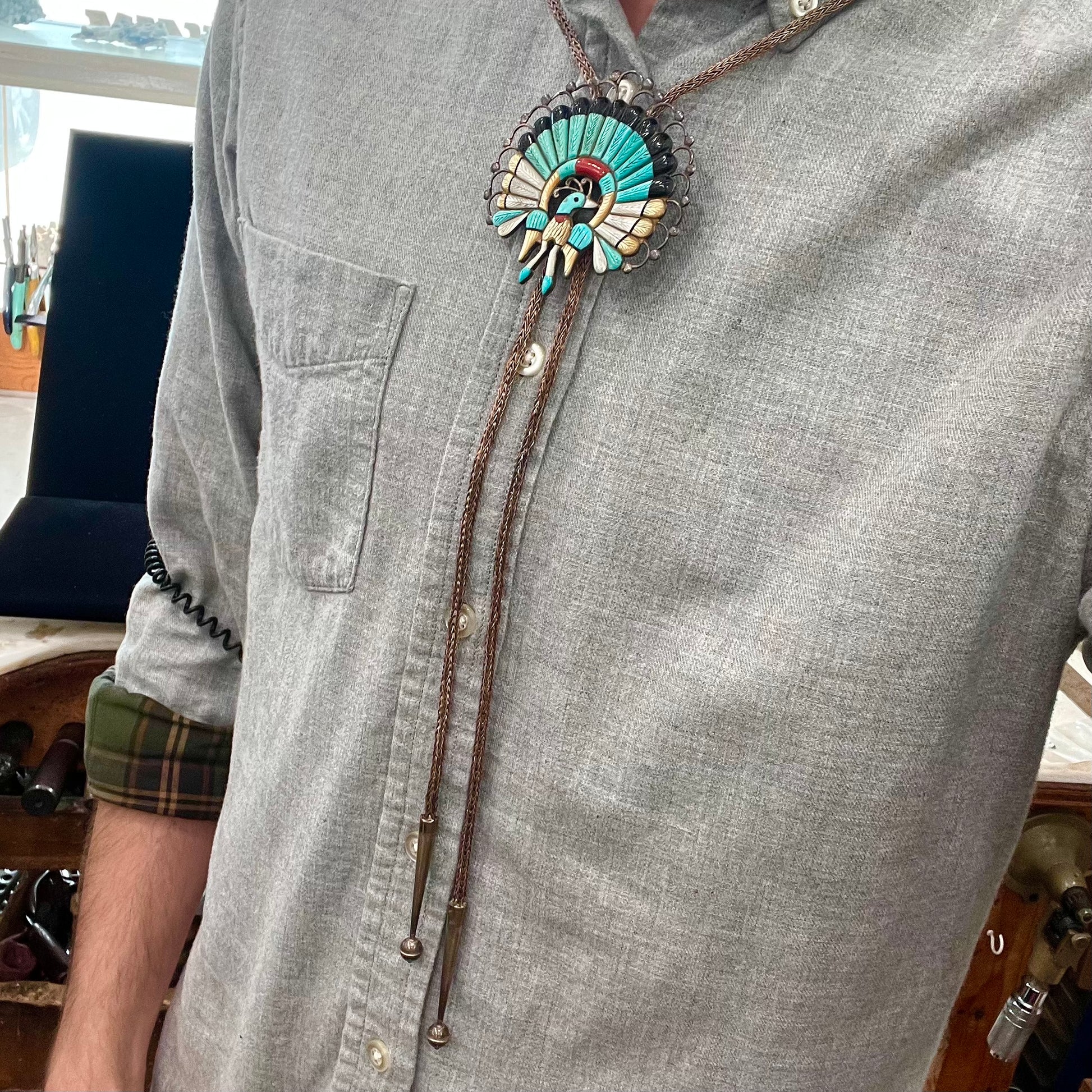 A handmade Zuni style stone inlay peacock bolo tie set with onyx, coral, turquoise, and gold lipped pearl on a silver bolo chain.