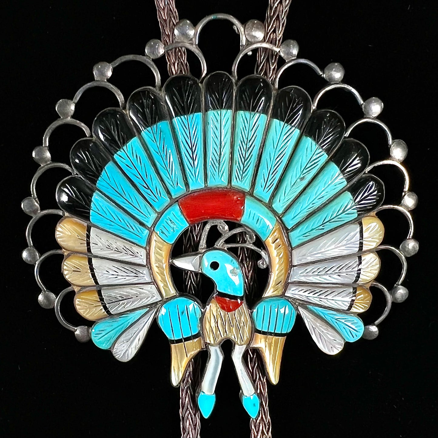 A handmade Zuni style stone inlay peacock bolo tie set with onyx, coral, turquoise, and gold lipped pearl on a silver bolo chain.