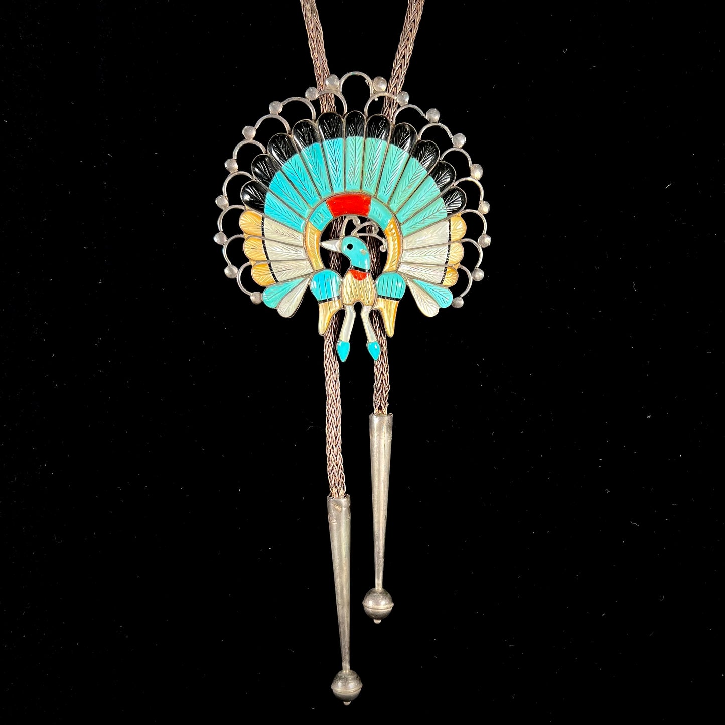 A handmade Zuni style stone inlay peacock bolo tie set with onyx, coral, turquoise, and gold lipped pearl on a silver bolo chain.