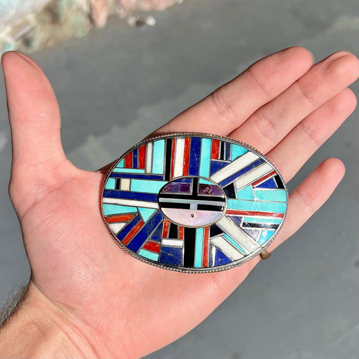 A sterling silver Zuni sunface belt buckle inlaid with lapis, turquoise, onyx, mother of pearl, coral, and sugilite stones.  It is stamped "RGB Zuni."